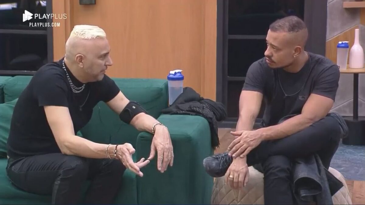 Rogério sentado no sofá verde na sala do Power Couple de frente para Mussunzinho de pernas cruzadas.