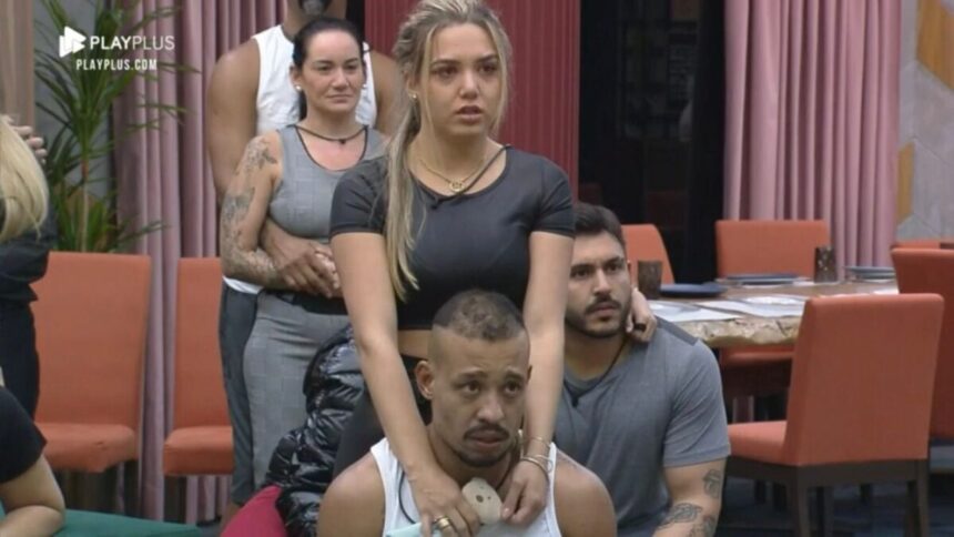 Karol em pé com cara de nervosa e Mussunzinho sentado próximo a esposa.