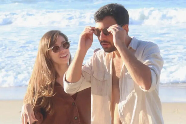 Casal Dani de óculos de sol com camisa marrom e cabelo solto e Luiz ajeitando o óculos, de camisa social branca, na praia durante ensaio fotográfico da gravação do clipe de "Viver"