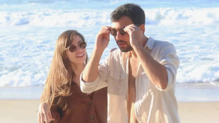 Casal Dani de óculos de sol com camisa marrom e cabelo solto e Luiz ajeitando o óculos, de camisa social branca, na praia durante ensaio fotográfico da gravação do clipe de "Viver"