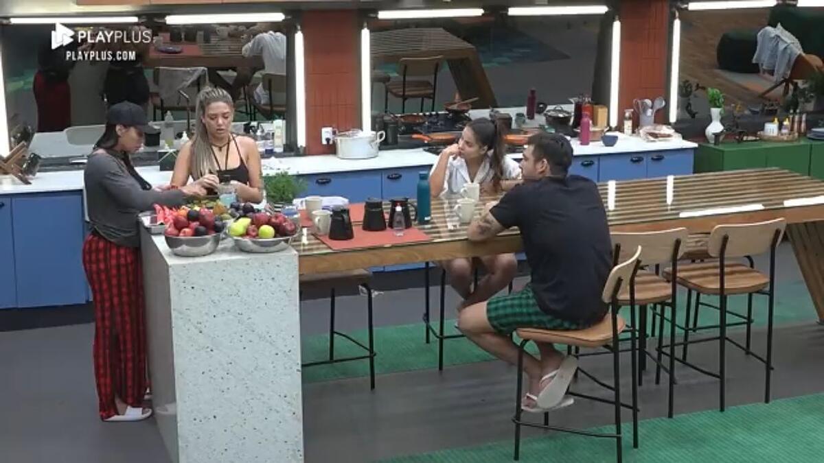 Eliza em pé no canto da mesa do Power couple ao lado de Karol. Em outra parte da mesa, Luana e Hadad sentados um de frente do outro.