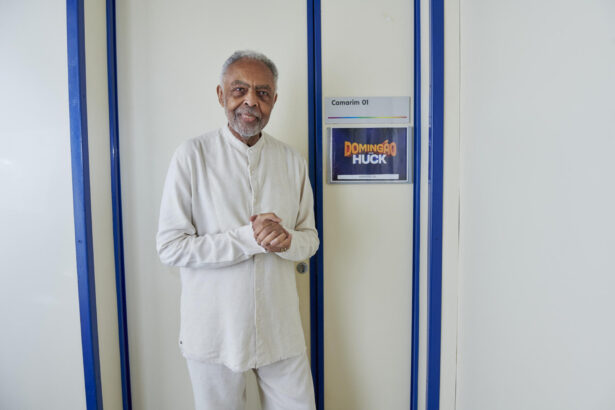 Gilberto Gil de roupa toda branca nos bastidores do Domingão. Ele posa em frente ao camarim com o logo do programa