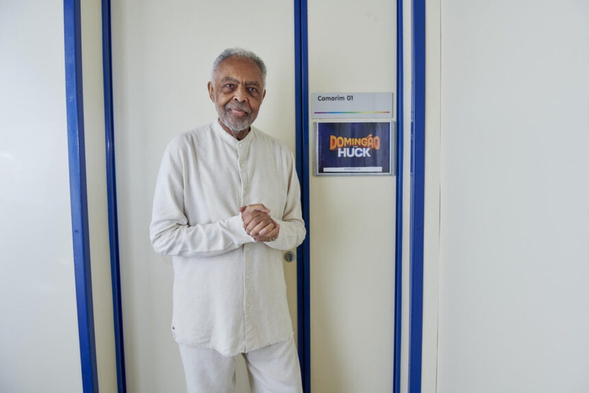 Gilberto Gil de roupa toda branca nos bastidores do Domingão. Ele posa em frente ao camarim com o logo do programa