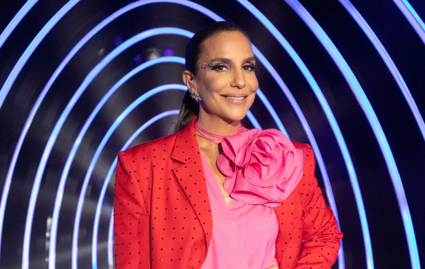 Ivete Sangalo com uma blusa rosa e um flor em cima do busto, nos estúdios do The Masked Singer Brasil