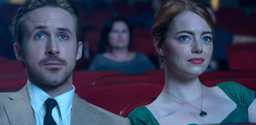 Ryan Gosling de palitó e gravata e Emma Stone de vestido verde e batom vermelho olhando para a tela do cinema durante gravação do filme La La Lan - Cantando Estações