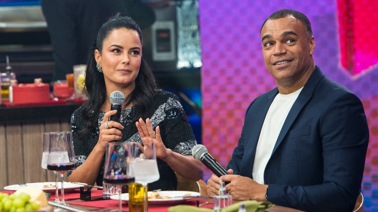 Luciele di Camargo de vestido preto, com o microfone na mão olhando para frente; Denilson de blazer azul e camisa básica branca com o microfone na mão olhando pra cima na gravação do Faustão da Band, da Band