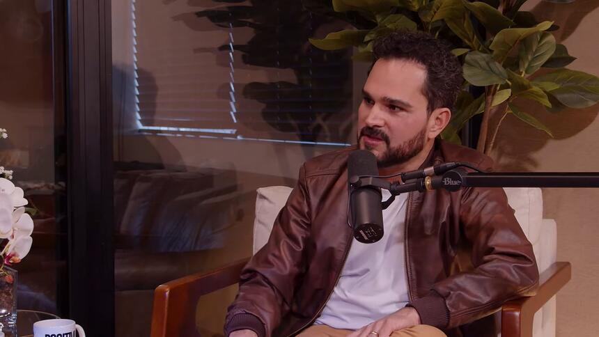 Luciano Camargo em trecho da entrevista para o podcast, com um microfone de rádio próximo ao seu rosto