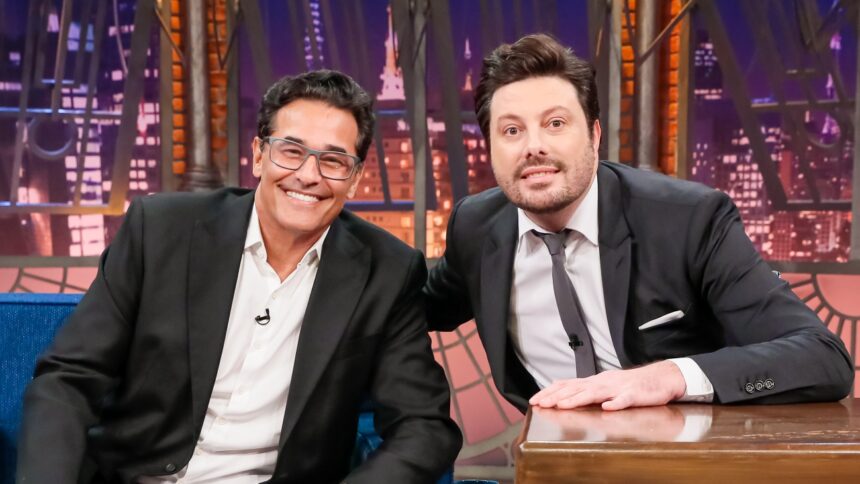Luciano Szafir está de óculo de grau, palitó preto e camisa social branca, sentado num sofá azul, ao lado de Danilo Gentili, de palitó e gravata preta, na bancada do The Noite, durante gravação