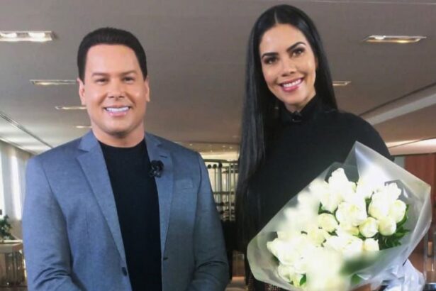 Marcos Paulo Ribeiro, o Marcão do Povo, de blaser cinza e camisa preta, sorrindo, ao lado de Daniela Albuquerque, de vestido preto, cabelo liso, solto, sorrindo, segurando um buquê de flores durante gravação de entrevista para o Sensacional, da RedeTV!