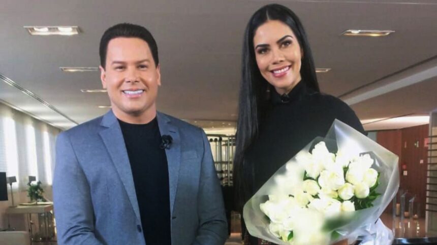 Marcos Paulo Ribeiro, o Marcão do Povo, de blaser cinza e camisa preta, sorrindo, ao lado de Daniela Albuquerque, de vestido preto, cabelo liso, solto, sorrindo, segurando um buquê de flores durante gravação de entrevista para o Sensacional, da RedeTV!