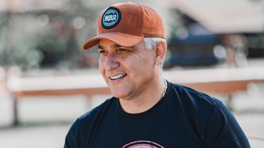 Locutor Marco Brasil de chapéu marron e camisa preta, na fazenda, sorrindo e olhando pro lado