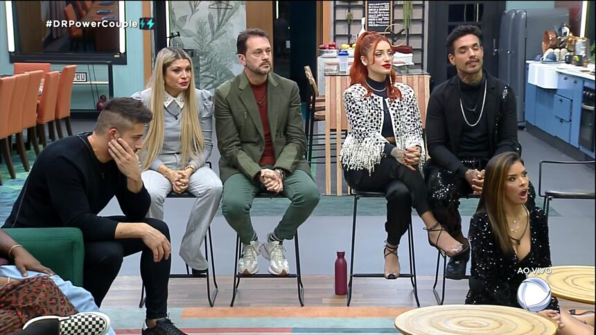 Nandinho, Adryana, Albert, Brenda, Matheus e Ivy assistem ao vídeo na TV da sala do Power couple que mostra cochicho de Anne e Karol