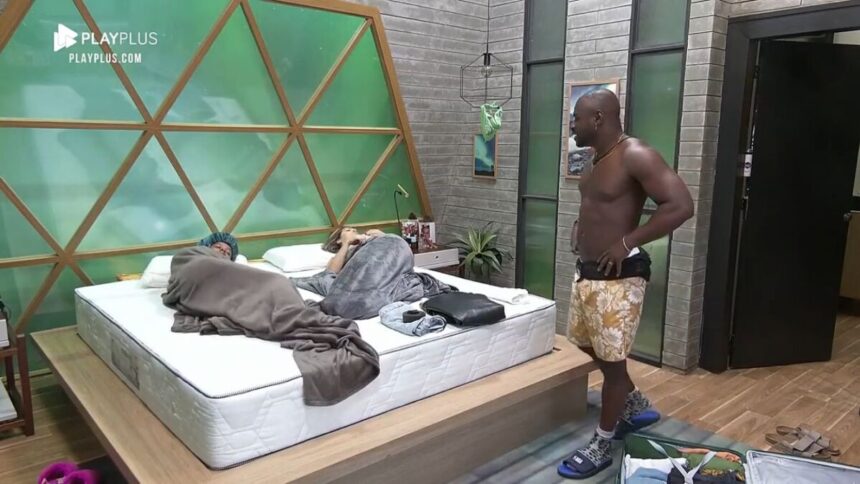 Bruno Passa em pé no quarto do Power Couple. Michele e Karol enroladas na cama.