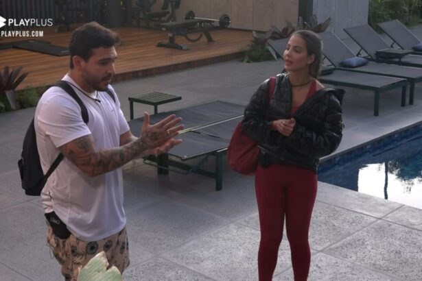 Hadad de camisa branca e bermuda estampada ao lado de Luana que segura uma bolsa na área externa do Power Couple.