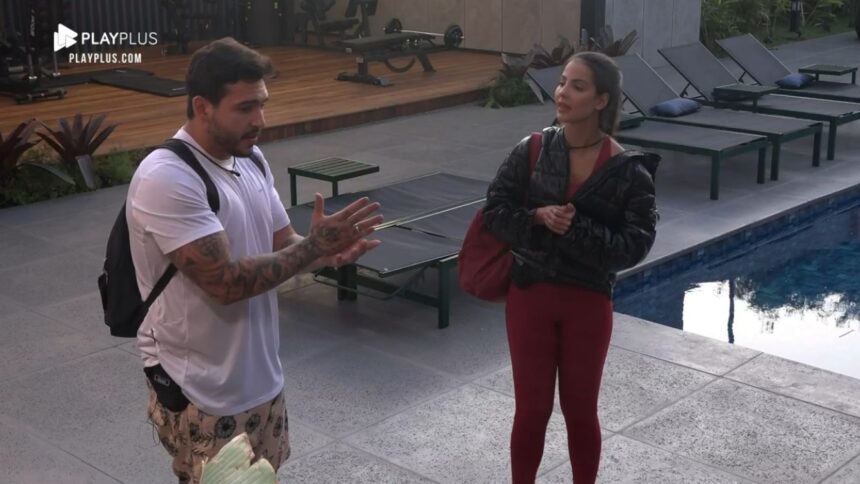 Hadad de camisa branca e bermuda estampada ao lado de Luana que segura uma bolsa na área externa do Power Couple.