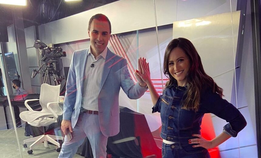 Rafael Colombo e Marcela Rahal separados por um vidro nos estúdios da CNN Brasil. Eles estão juntando as mãos