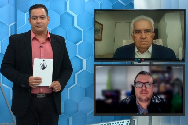 Jornalista Eduardo Miranda em pé, de palitó e camisa rosa, segurando um folheto ao lado de dois monitores com exibição de chamada de video com Raul Velloso e Luiz Henrique Mourão