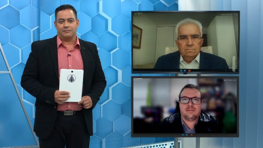 Jornalista Eduardo Miranda em pé, de palitó e camisa rosa, segurando um folheto ao lado de dois monitores com exibição de chamada de video com Raul Velloso e Luiz Henrique Mourão