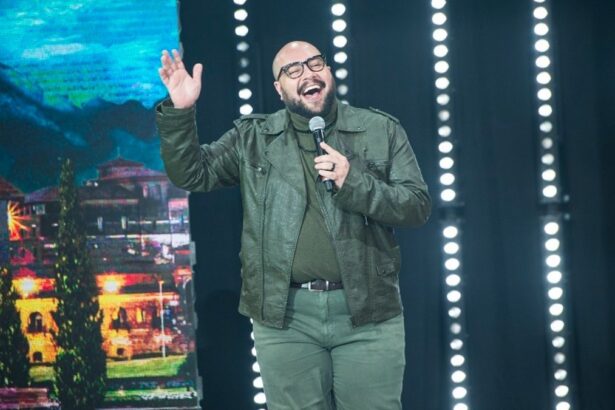 Tiago Abravanel de jaqueta, camisa e calça jeans cinza, com microfone na mão, acenando para a plateia durante gravação do Faustão na Band