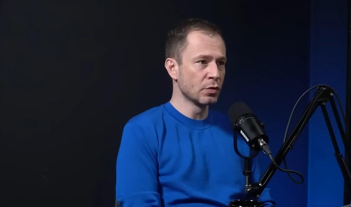 Tiago Leifert de blusa azul participando de um podcast. Um microfone de rádio em tripé a sua frente