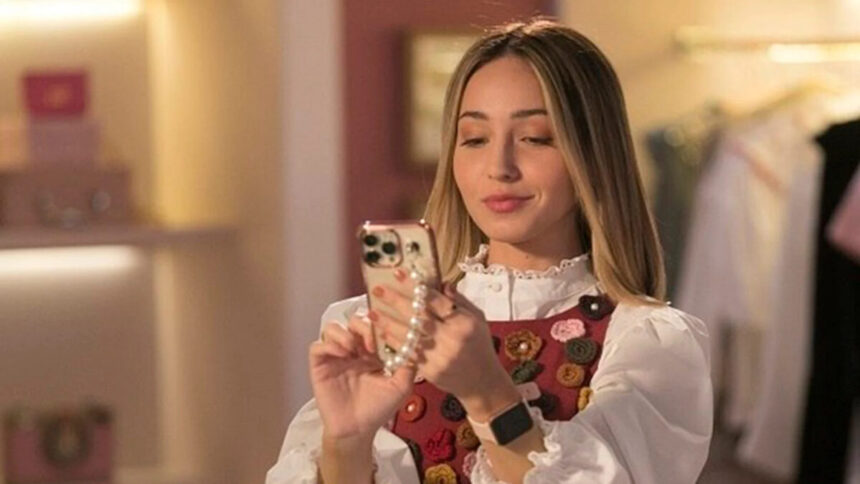 Atriz Mharessa Fernanda, de camisa mangalonga branca e sobreposição estampada com bolas coloridas, mexendo no celular, durante gravação da série Todas as Garotas em Mim (Record TV)