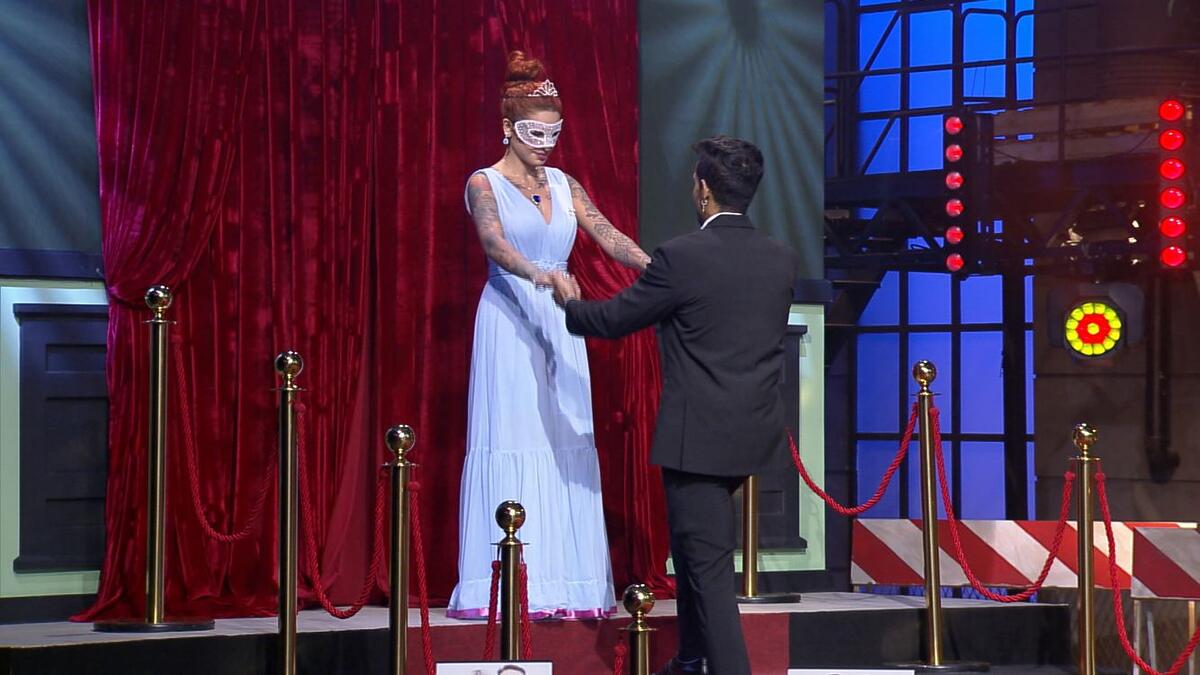 Brenda vestida com um vestido longo branco e de máscara. Na sua frente, Matheus vestido de roupa de gala.