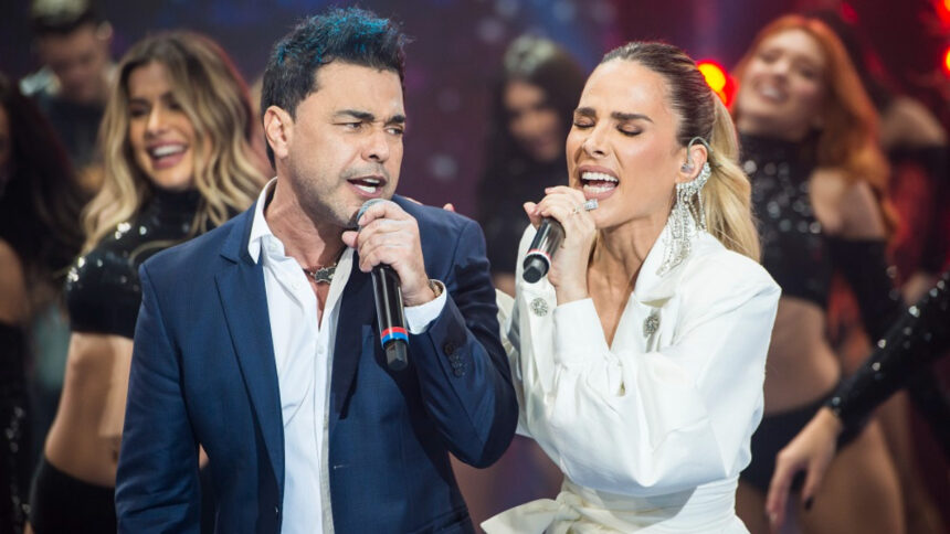 Zezé di Camargo de blazer azul e camisa social branca, com o microfone na mão esquerda ao lado de Wanessa, de camisa social branca, com microfone na mão esquerda durante gravação do Faustão na Band, da Band