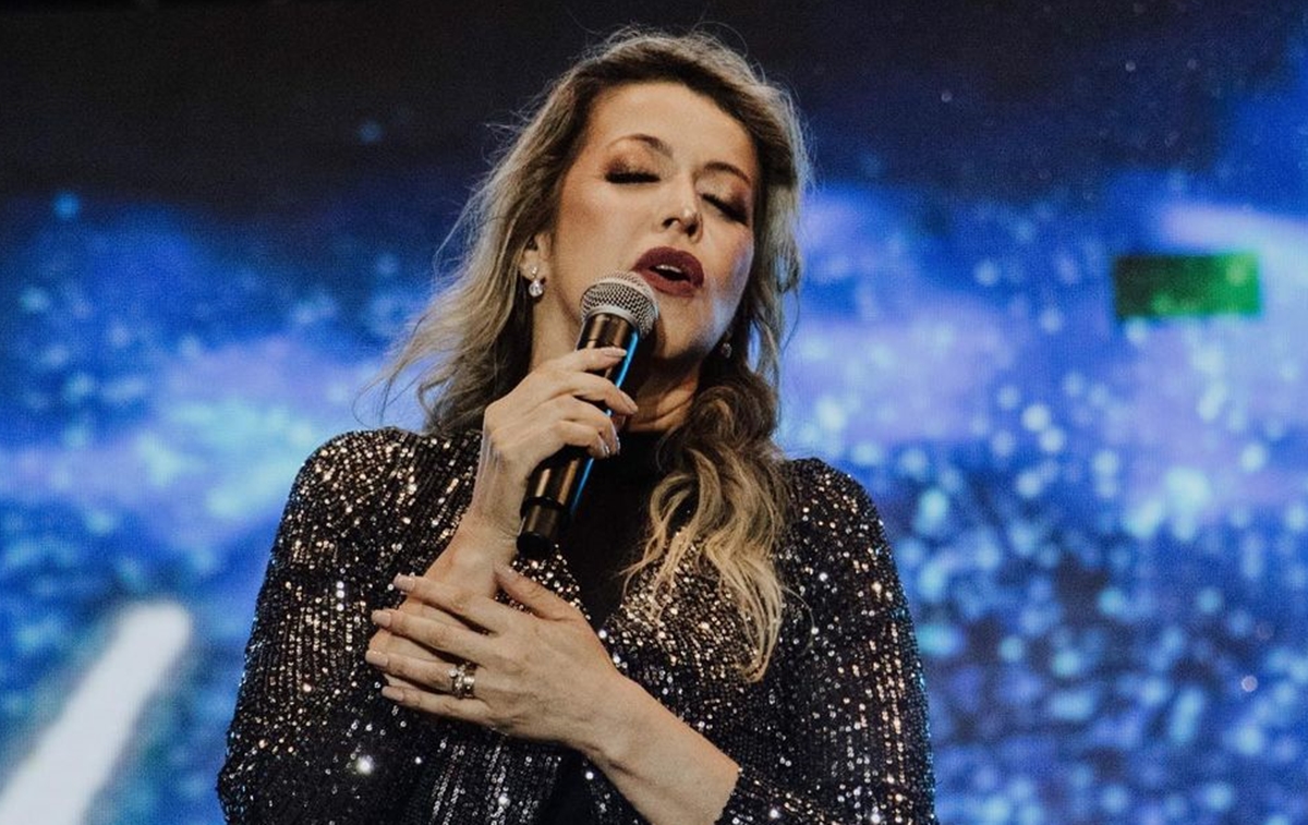 Adriana Arydes com camisa manga longa preta, segurando o microfone com a mão direita, durante apresentação em show