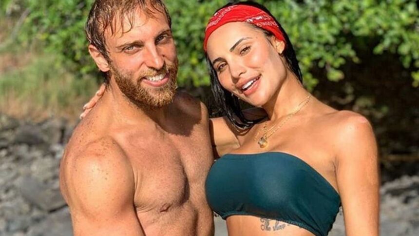 Leo Lins sorrindo, sem camisa, abraçando Aline Mineiro, que está sorrindo, de biquine verde e bandana na cabeça