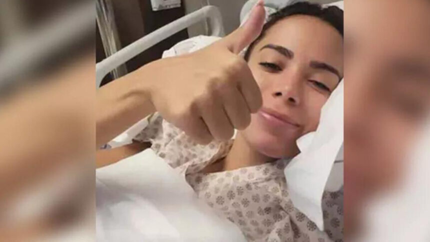 Anitta faz o sinal de positivo com a mão, cantora está deitada em um leito de hispital