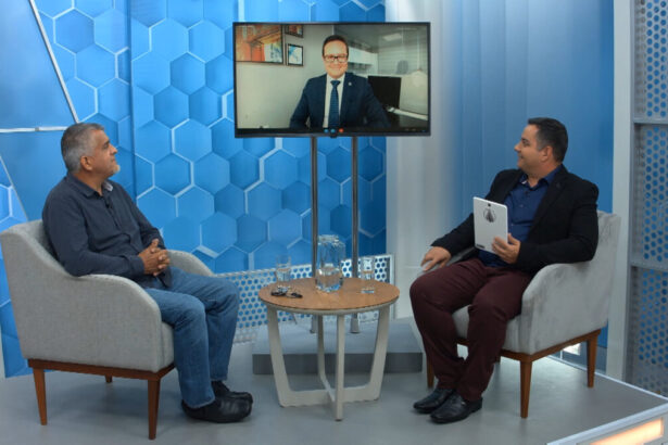 Jornalista Renato Dias de blazer preto, camisa azul e calça preta segurando as fichas do programa; olhando para o monitor, dialogando com um dos convidados com expressão séria; concentrado no debate