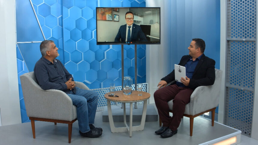 Jornalista Renato Dias de blazer preto, camisa azul e calça preta segurando as fichas do programa; olhando para o monitor, dialogando com um dos convidados com expressão séria; concentrado no debate