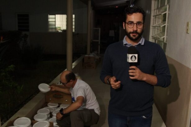 Guilherme Machado segurando um microfone com a mão esquerda, de camisa de frio azul escura, de óculos, durante gravação acompanhando uma ação beneficente