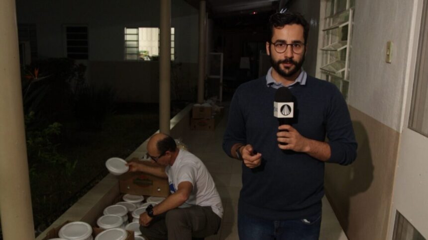 Guilherme Machado segurando um microfone com a mão esquerda, de camisa de frio azul escura, de óculos, durante gravação acompanhando uma ação beneficente