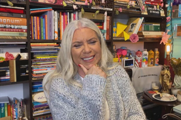 Astrid Fontenelle em sua biblioteca, sorrindo, com camisa manga-longa cinza, com a mão no queixo durante entrevista a Pedro Bial