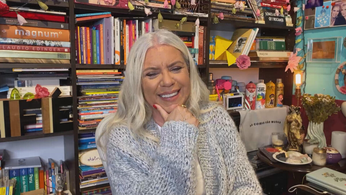 Astrid Fontenelle em sua biblioteca, sorrindo, com camisa manga-longa cinza, com a mão no queixo durante entrevista a Pedro Bial