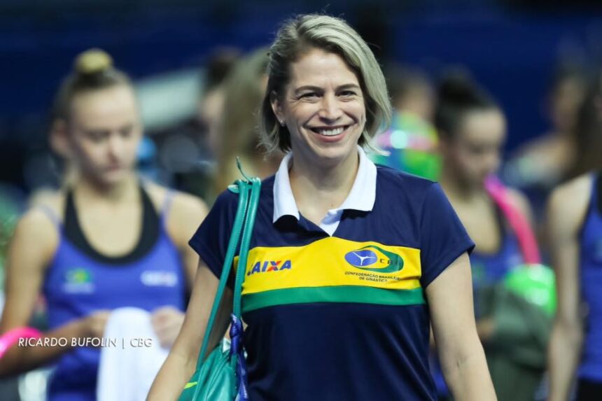 Camila Ferezin com a camisa de treinadora da Seleção Brasileira nos Jogos Olímpicos, sorrindo, com uma bolsa verde no ombro direito