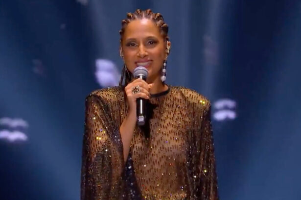 Camila Pitanga com vestido em transparência e brilhoso na cor preta, segurando um microfone com a mão direita e penteado em tranças durante apresentação de homenagem a Elza Soares