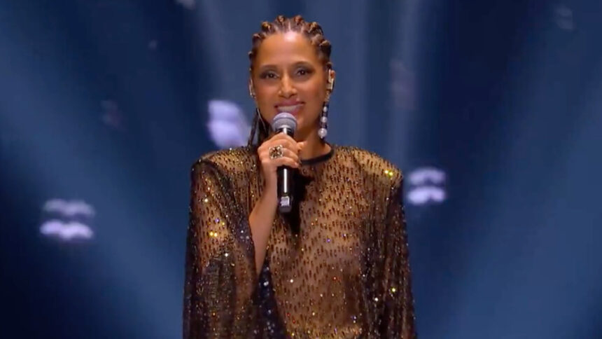 Camila Pitanga com vestido em transparência e brilhoso na cor preta, segurando um microfone com a mão direita e penteado em tranças durante apresentação de homenagem a Elza Soares