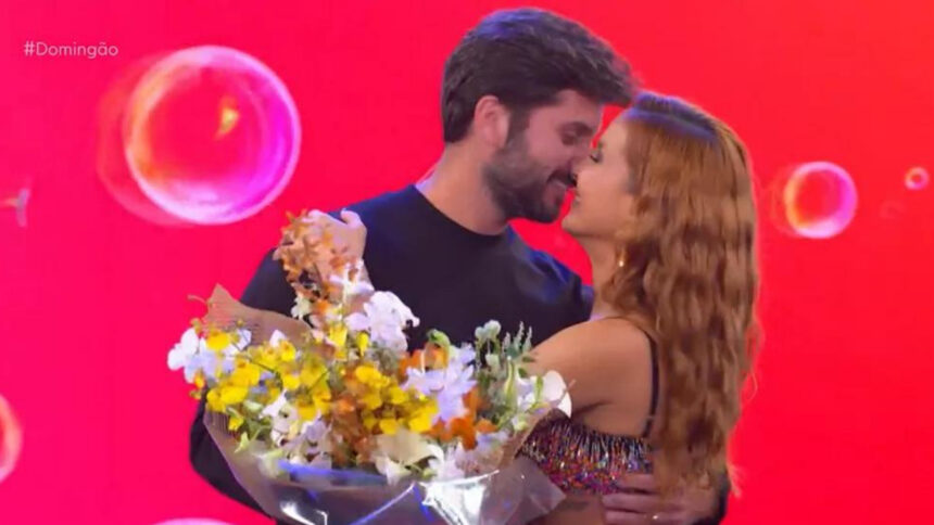 Claudia Leitte abraçada com o marido no palco do Domingão. Ele segura um buquê de flores com uma das mãos