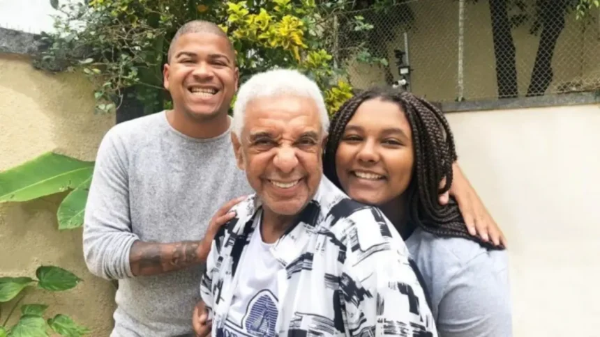 Damião de camisa cinza, segurando no ombro de Agnaldo Timoteo, que está de camisa branca estampada com detalhes pretos, sorrindo, ao lado da filha, Keyty, que está sorrindo, com penteado de tranças e camisa cinza