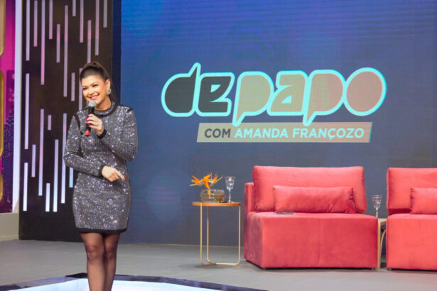 Amanda Françozo em pé, nos estúdios do De Papo com Amanda Françozo. Ao fundo, um telão e dois sofás dos convidados