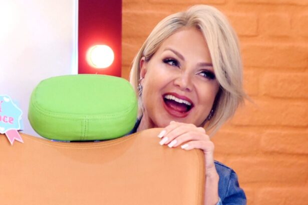 Eliana com a boca aberta de macacão azul atrás de um banco, segurando o microfone com a mão direita durante gravação do seu programa no SBT