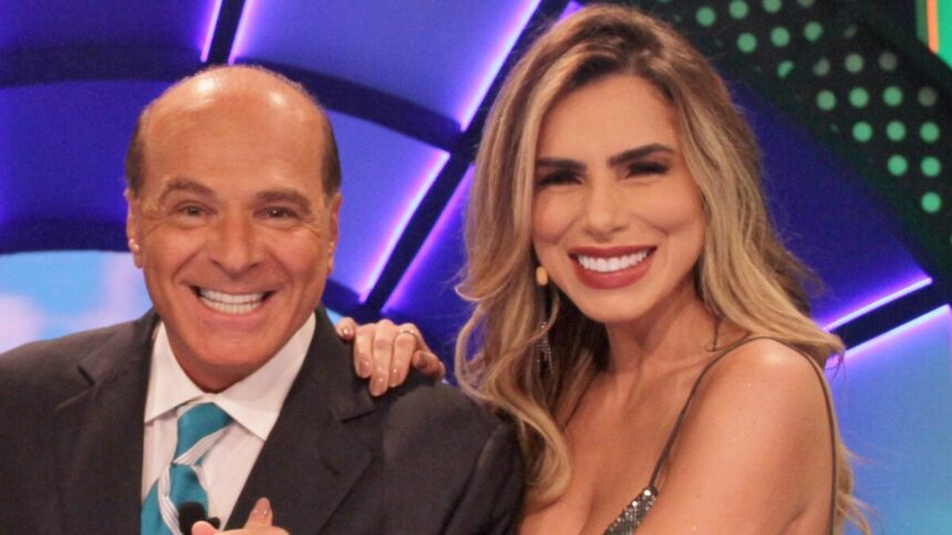 Marcelo de Carvalho de palitó preto, camisa social branca e gravata azul ao lado de Erika Schneider, sorrindo, de microfone no rosto e vestido prata durante gravação do "O Céu é o Limite", da RedeTV!