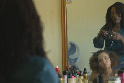 Repórter Daniella Dias fazendo um trança afro, em um salão de beleza em frente a um espelho, em trecho do Expedição Rio