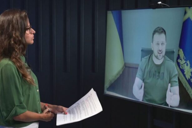 Raquel Krähenbühl em pé, de óculos, com camisa verde, segurando papéis em frente a um monitor conversando com o presidente da Ucrânia, Volodymyr Zelensky, para o Fantástico, da TV Globo