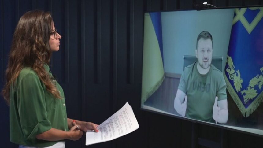 Raquel Krähenbühl em pé, de óculos, com camisa verde, segurando papéis em frente a um monitor conversando com o presidente da Ucrânia, Volodymyr Zelensky, para o Fantástico, da TV Globo