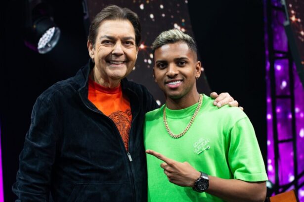 Apresentador Fausto Silva de blusão preto e camisa laranja, sorrindo, com a mão no ombro de Rodrygo Goes, sorrindo, de colar de ouro, camisa verde apontando para Fasutão, durante gravação do Faustão na Band, da Band