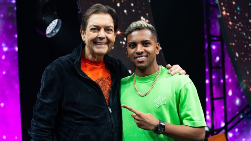 Apresentador Fausto Silva de blusão preto e camisa laranja, sorrindo, com a mão no ombro de Rodrygo Goes, sorrindo, de colar de ouro, camisa verde apontando para Fasutão, durante gravação do Faustão na Band, da Band