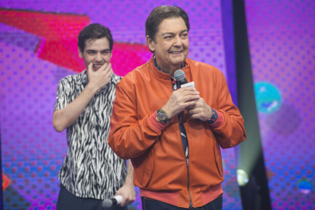 Faustão de camisa laranja, sorrindo, segurando o microfone; João Guilherme, seu filho, está atrás do apresentador, de camisa estampada, com a mão na boca, durante gravação